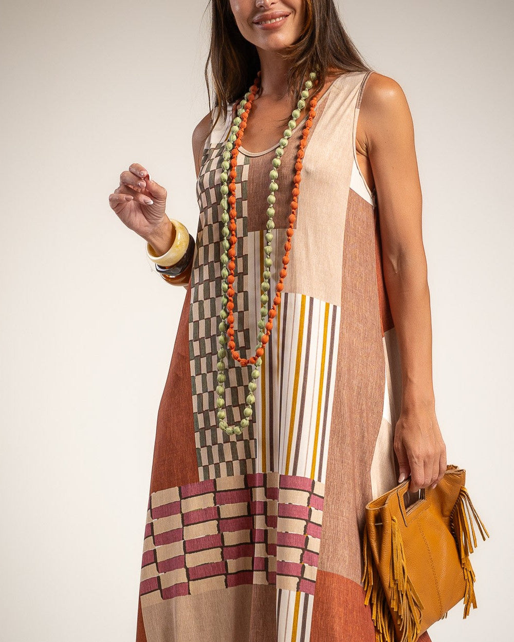 A woman wearing a sleeveless patchwork maxi dress with beige, brown, and rust-orange blocks, striped details, and a geometric pattern near the hem.