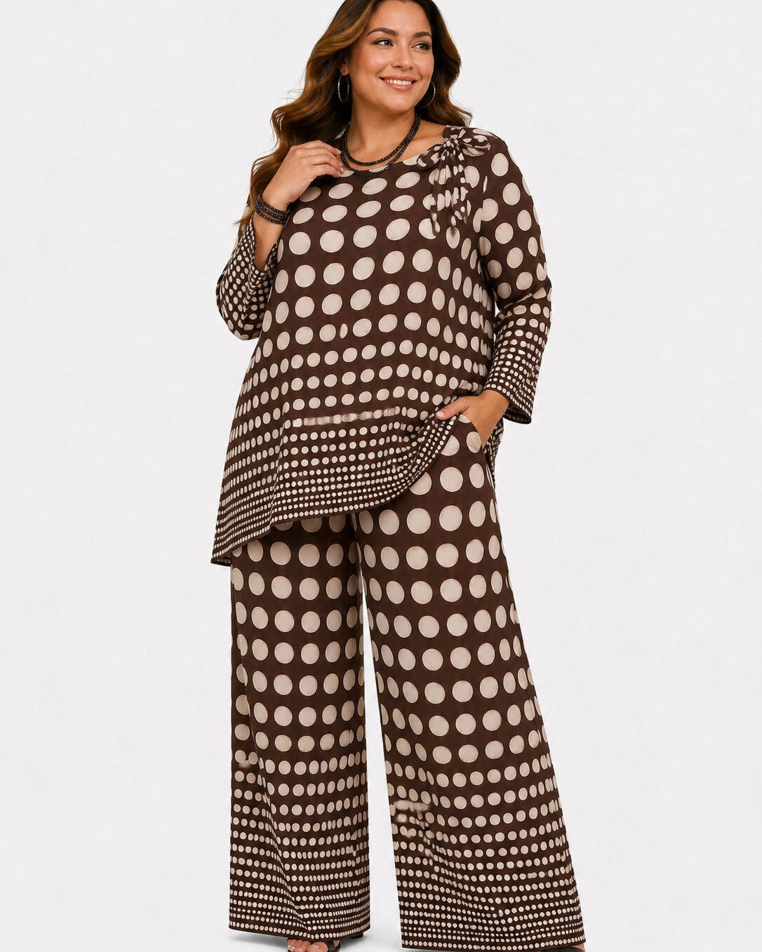 A woman modeling a matching two-piece outfit featuring a brown and beige polka dot pattern, consisting of a long-sleeve ruffled blouse and wide-leg pants, against a plain light gray background.