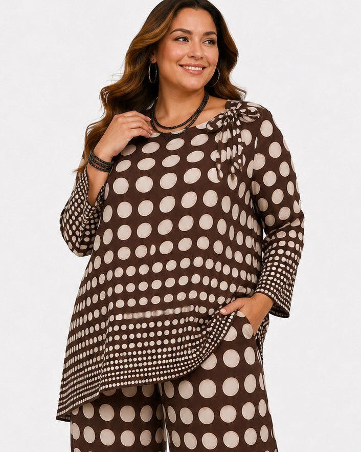A woman modeling a matching two-piece outfit featuring a brown and beige polka dot pattern, consisting of a long-sleeve ruffled blouse and wide-leg pants, against a plain light gray background.