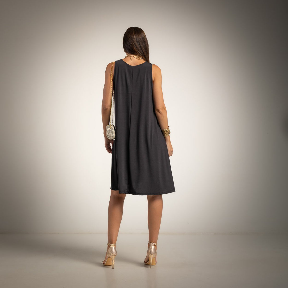 A woman is shown from the back wearing a sleeveless black A-line dress, paired with metallic gold high heels and a small white crossbody bag, set against a studio backdrop with a warm spotlight effect.