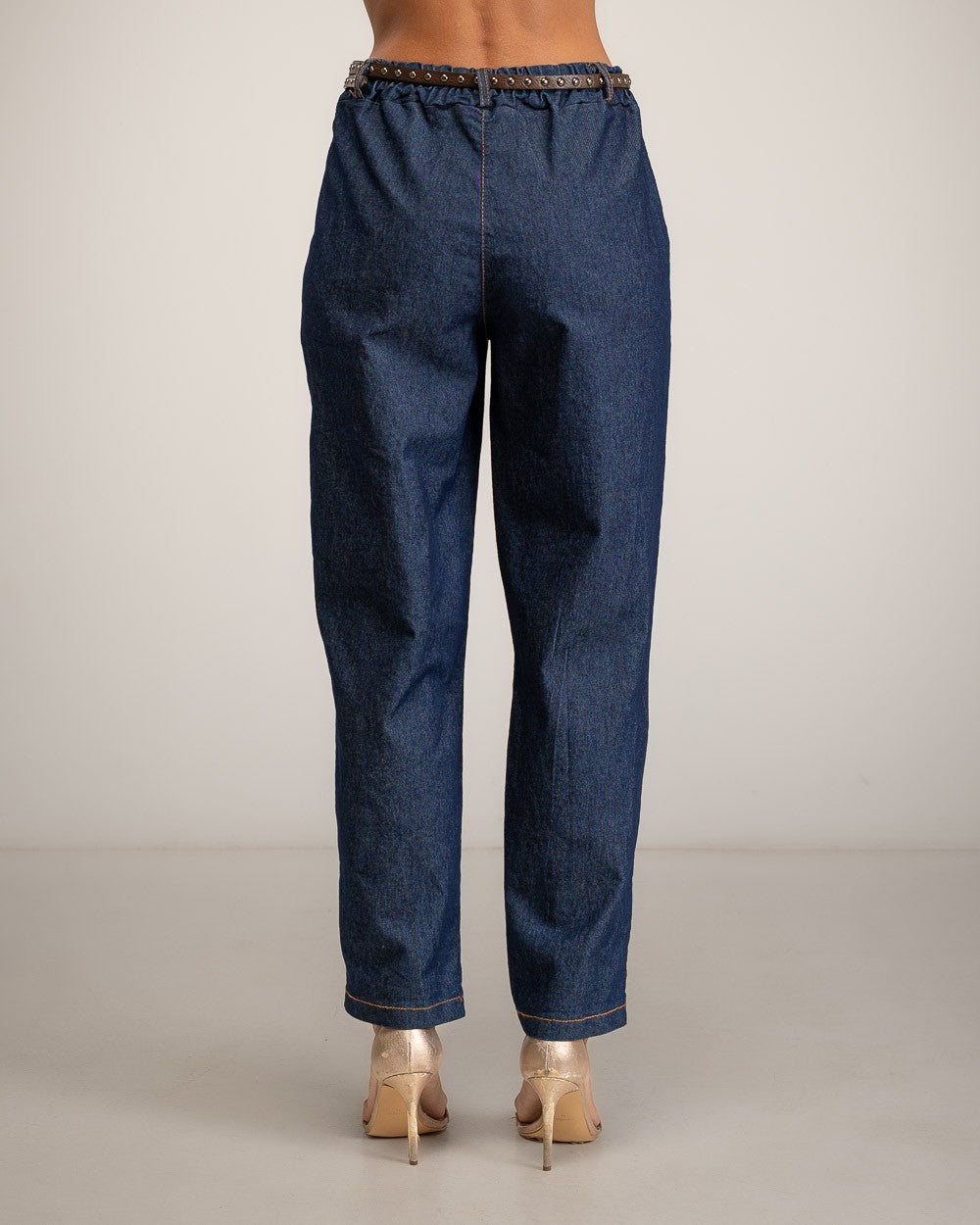 Rear view of a model wearing dark blue denim jeans with a decorative braided waistband and metallic gold high heels, set against a plain light gray studio background.