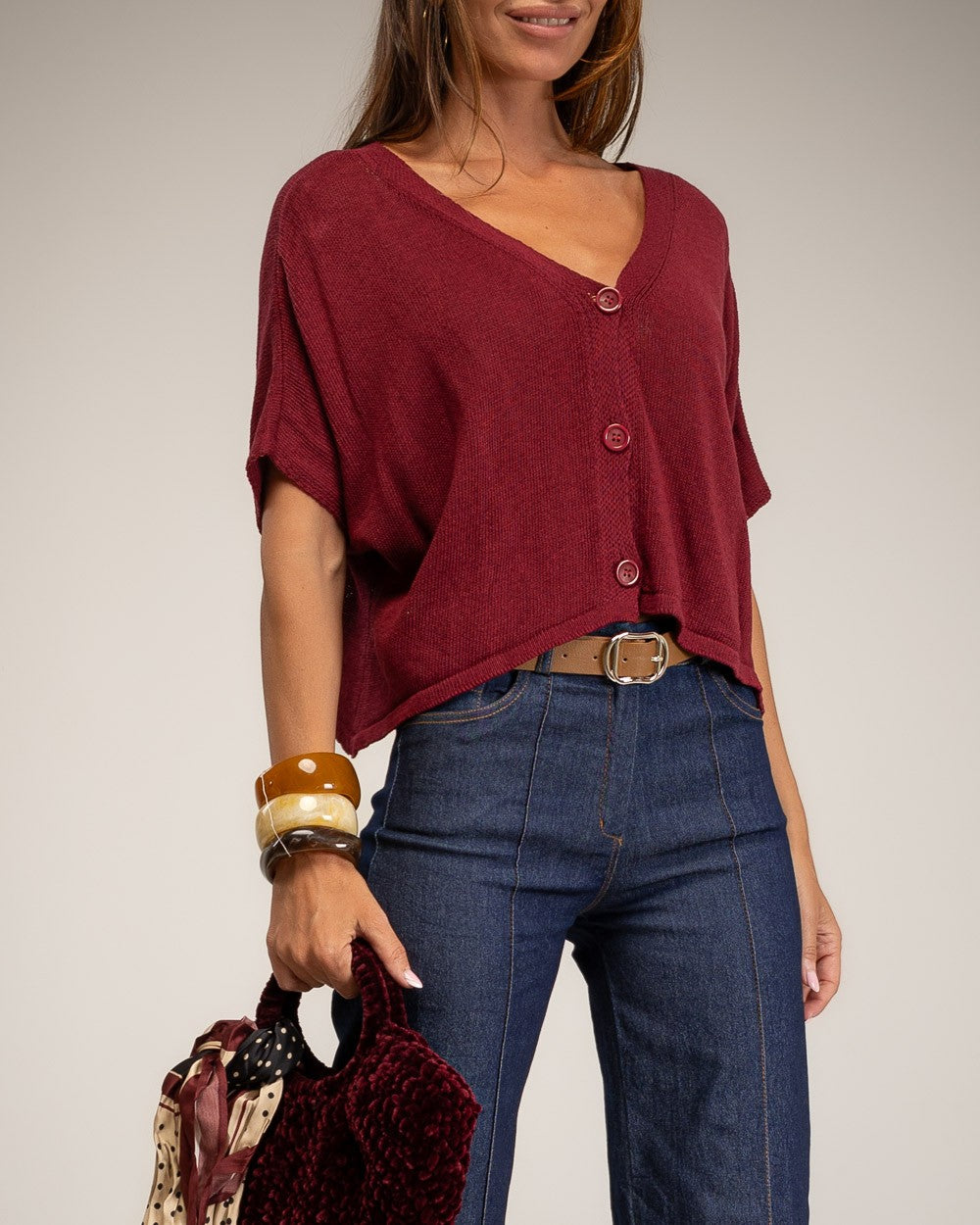 A woman wearing a burgundy short-sleeve button-front top with blue jeans, holding a patterned and velvet handbag against a plain light gray studio background.