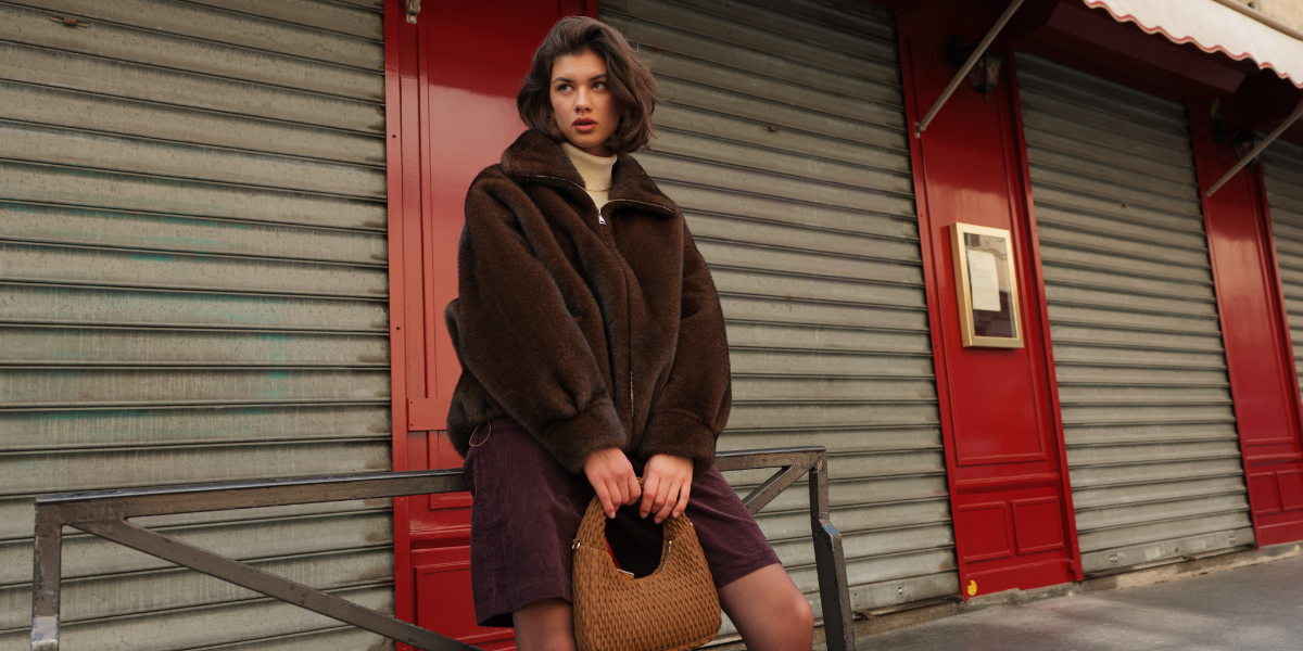 Woman in a brown coat standing in front of a red and gray shuttered door.