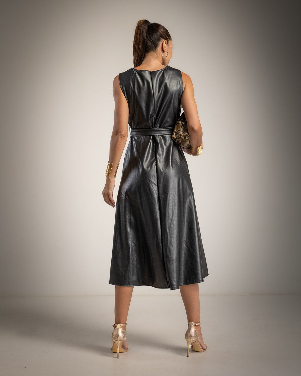 A woman shown from the back wearing a sleeveless black faux-leather midi dress with a matching belt, paired with gold high heels and holding a gold clutch, set against a neutral studio background.