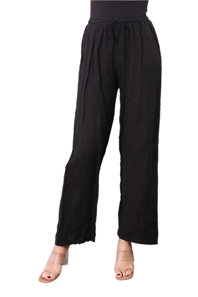 A woman standing against a white background wearing straight leg black trousers with an elasticated waist, paired with a black top. White background