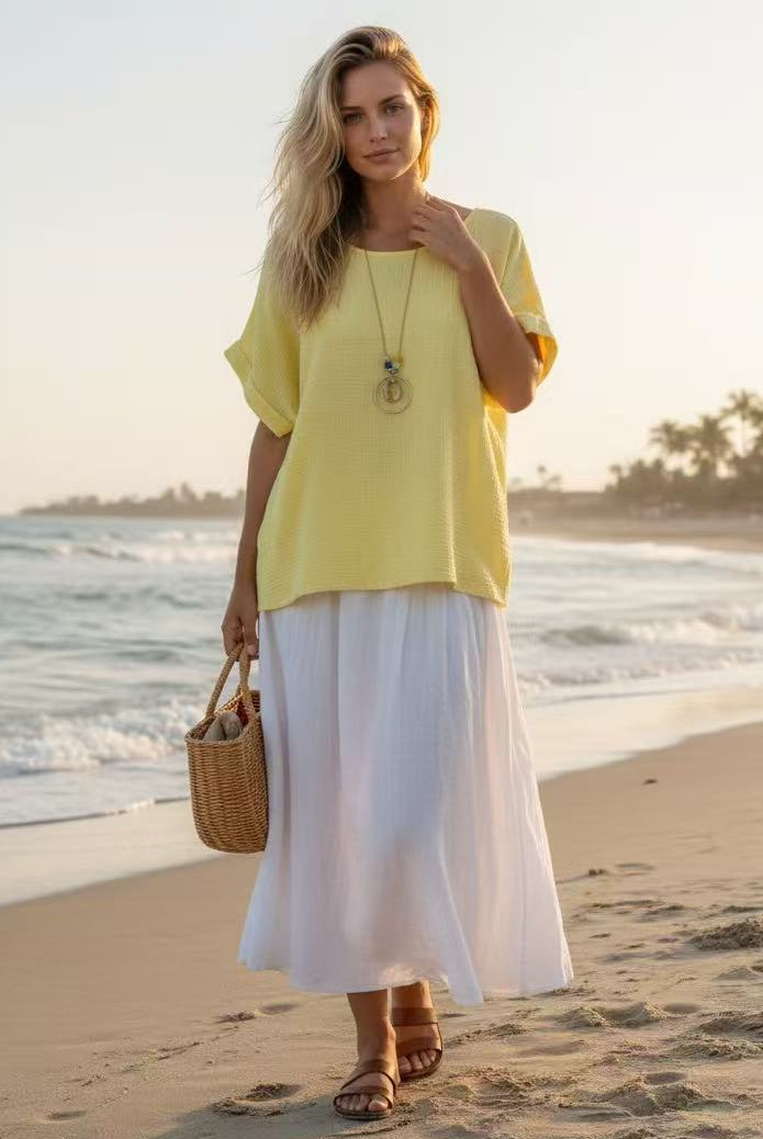 Woman in yellow top and white skirt standing on a beach with ocean and sunset in the background