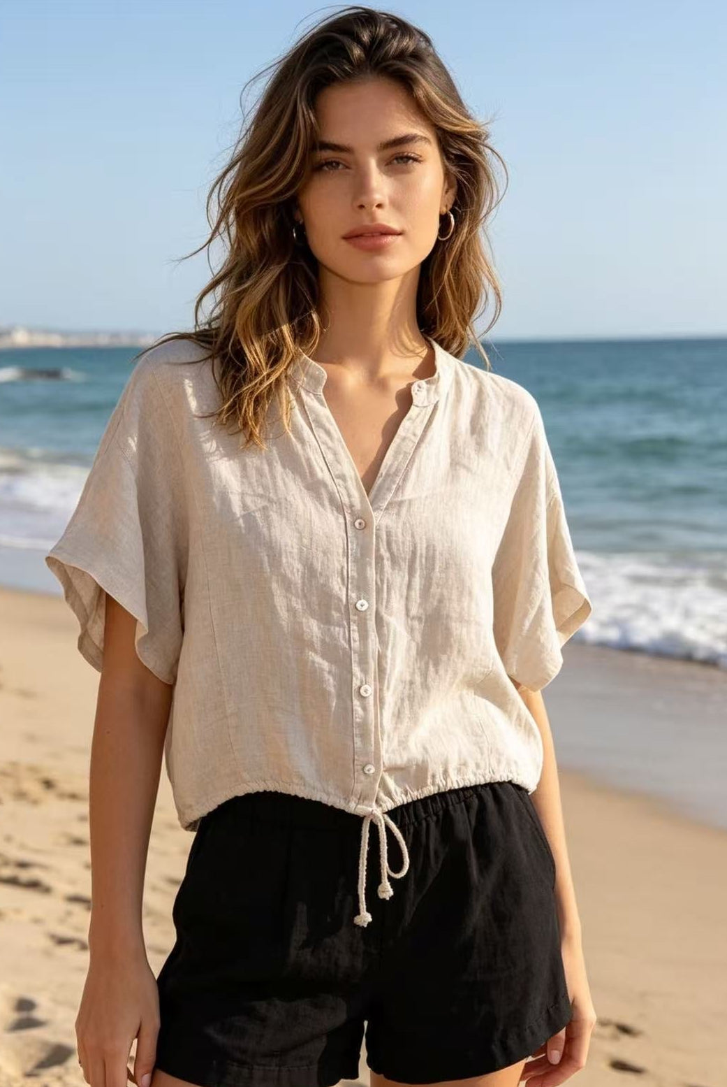 Woman wearing a light-colored shirt and black shorts on a beach with ocean in the background