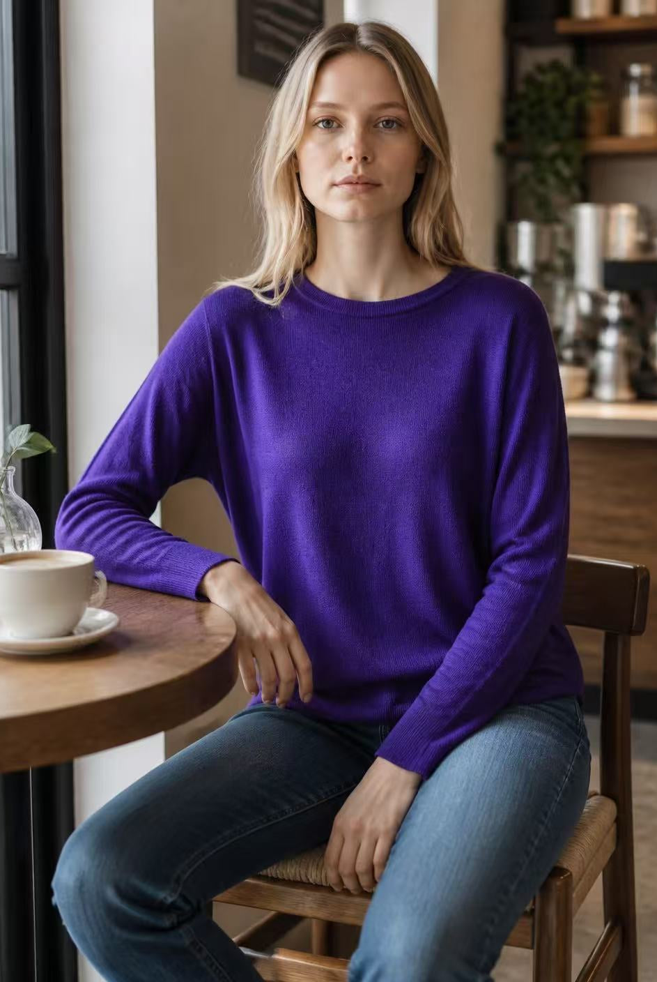 A woman with blonde hair sits on a wooden chair in a cozy cafe setting, wearing a vibrant purple long-sleeve crewneck sweater and blue jeans. A white coffee cup sits on a wooden table next to her.
