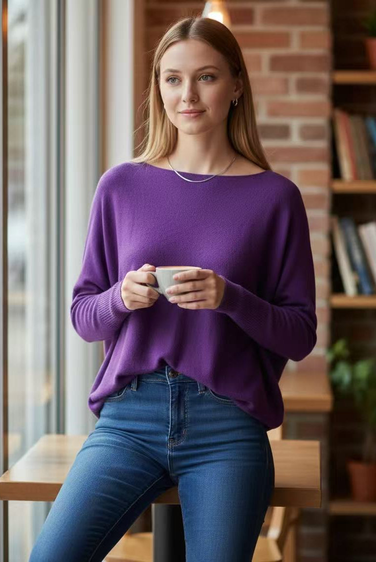 A woman wearing a purple long batwing sleeve knit sweater and blue jeans sits at a wooden table in a cozy indoor setting with a brick wall and bookshelves in the background, holding a white mug.