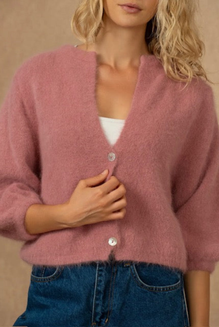 A woman wearing a fuzzy dusty pink cardigan with silver buttons over a white top and blue jeans, set against a warm beige background.