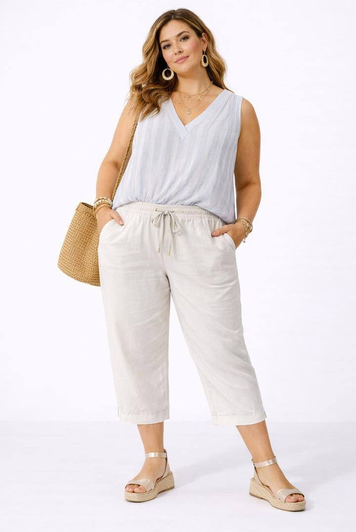 Woman wearing a white sleeveless top and beige pants, standing against a white background.