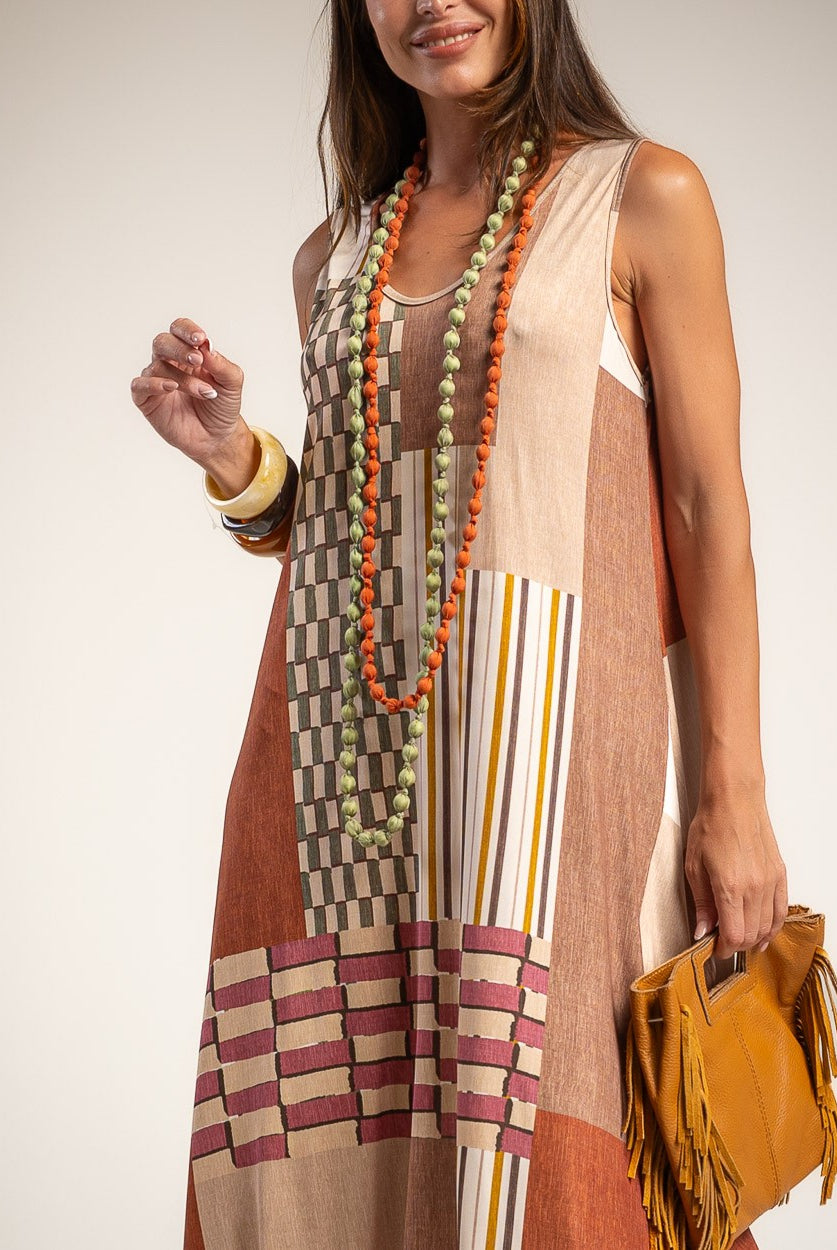 A woman wearing a sleeveless patchwork maxi dress with beige, brown, and rust-orange blocks, striped details, and a geometric pattern near the hem.