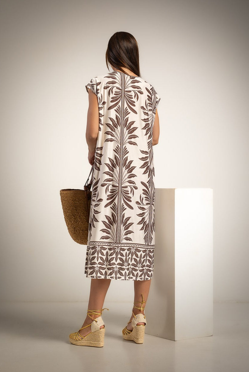 A woman stands with her back to the camera wearing a cream-colored dress with a brown botanical leaf pattern, holding a woven straw bag and standing next to a white display block in a studio setting.