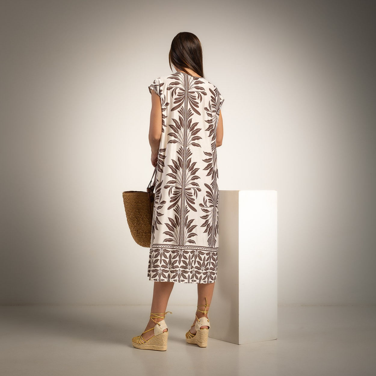A woman stands with her back to the camera wearing a cream-colored dress with a brown botanical leaf pattern, holding a woven straw bag and standing next to a white display block in a studio setting.