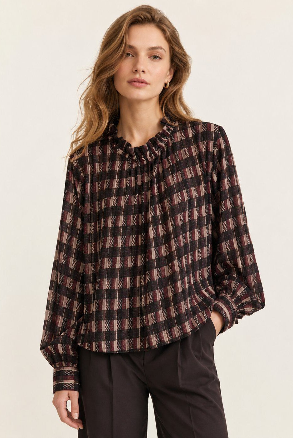 A woman wearing a long-sleeve plaid blouse with a gathered ruffled neckline, paired with dark trousers, against a plain light beige studio background.