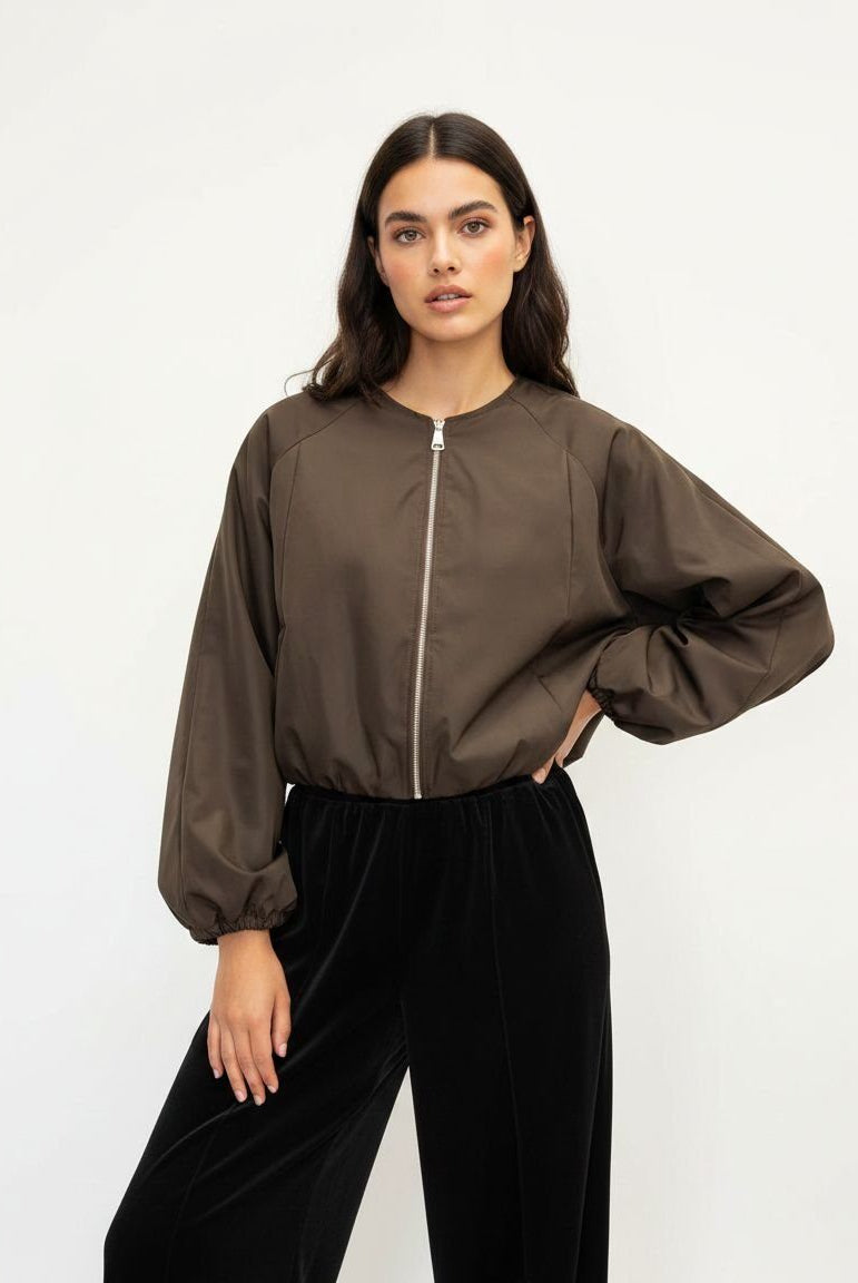 A woman modeling a cropped, long-sleeve bomber-style jacket in a dark brown, paired with black velvet wide-leg pants, against a plain white studio background.