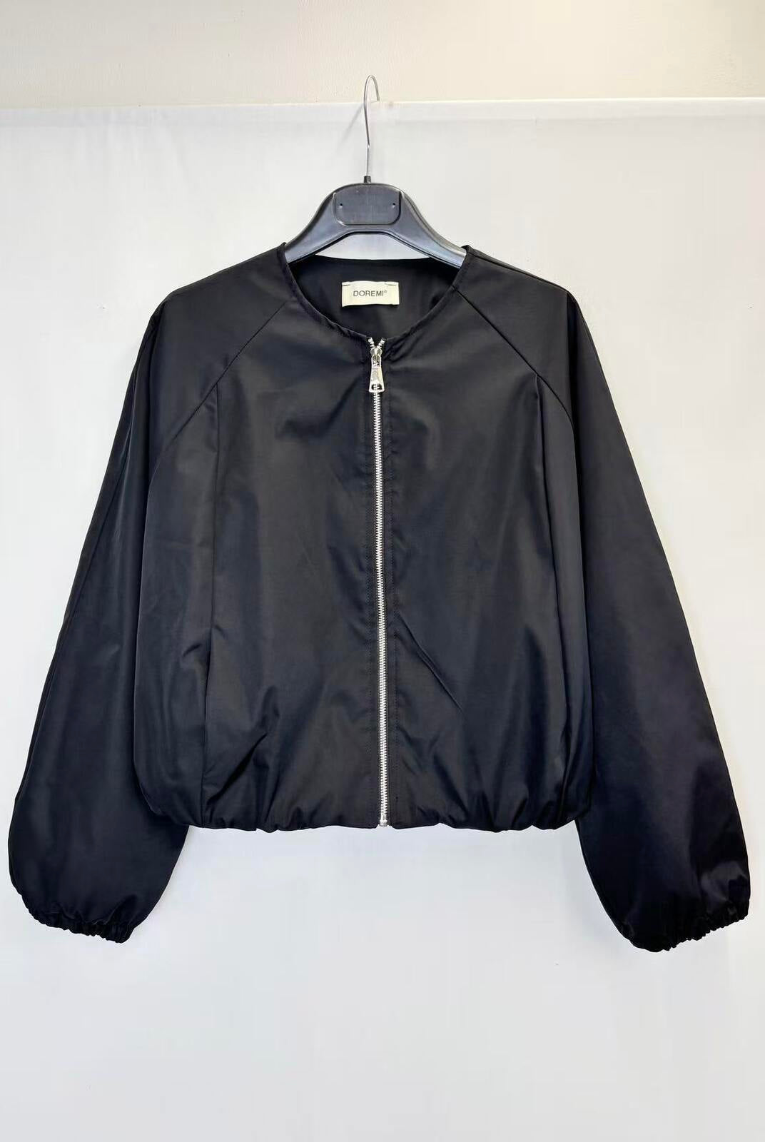 A black lightweight zip-up jacket hanging on a black hanger against a plain white wall. The jacket has a smooth, slightly shiny finish, elasticized cuffs and hem, and a silver zipper.