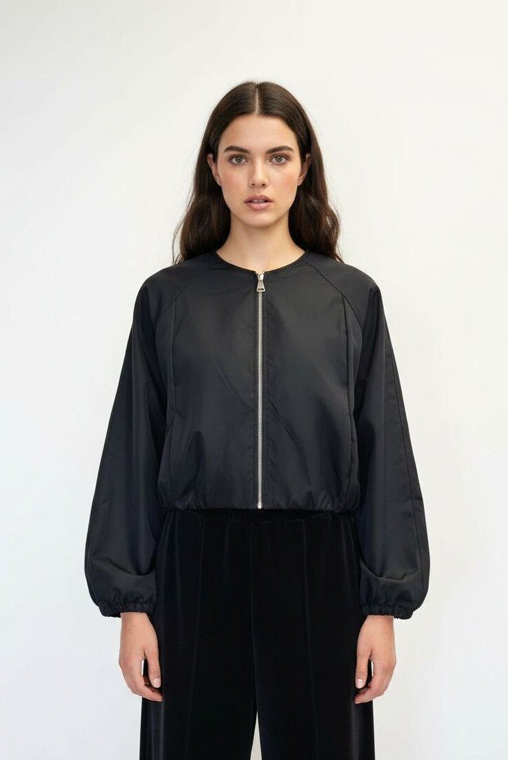 A woman modeling a black cropped bomber jacket with a silver front zipper and black wide-leg pants, shot against a plain white studio background.