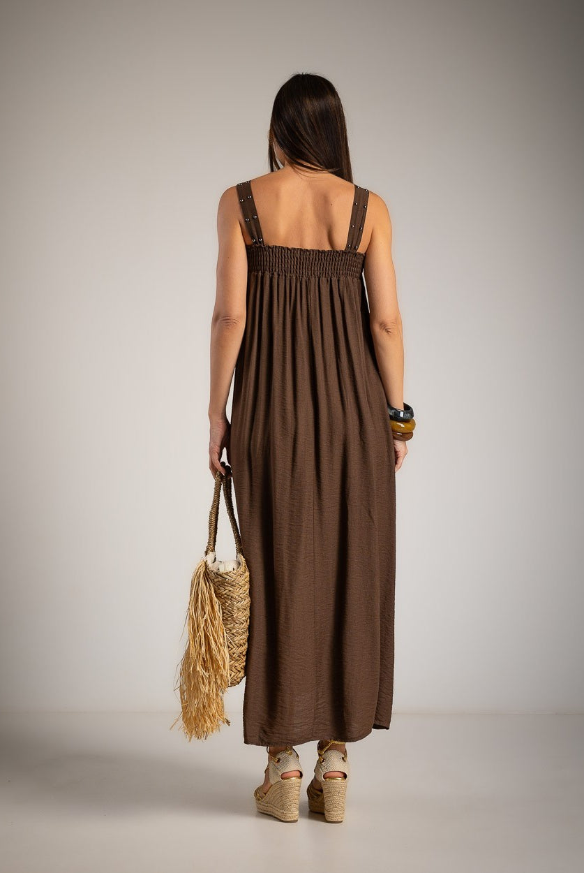 A woman viewed from behind wearing a sleeveless brown maxi dress with a smocked bodice and wide shoulder straps