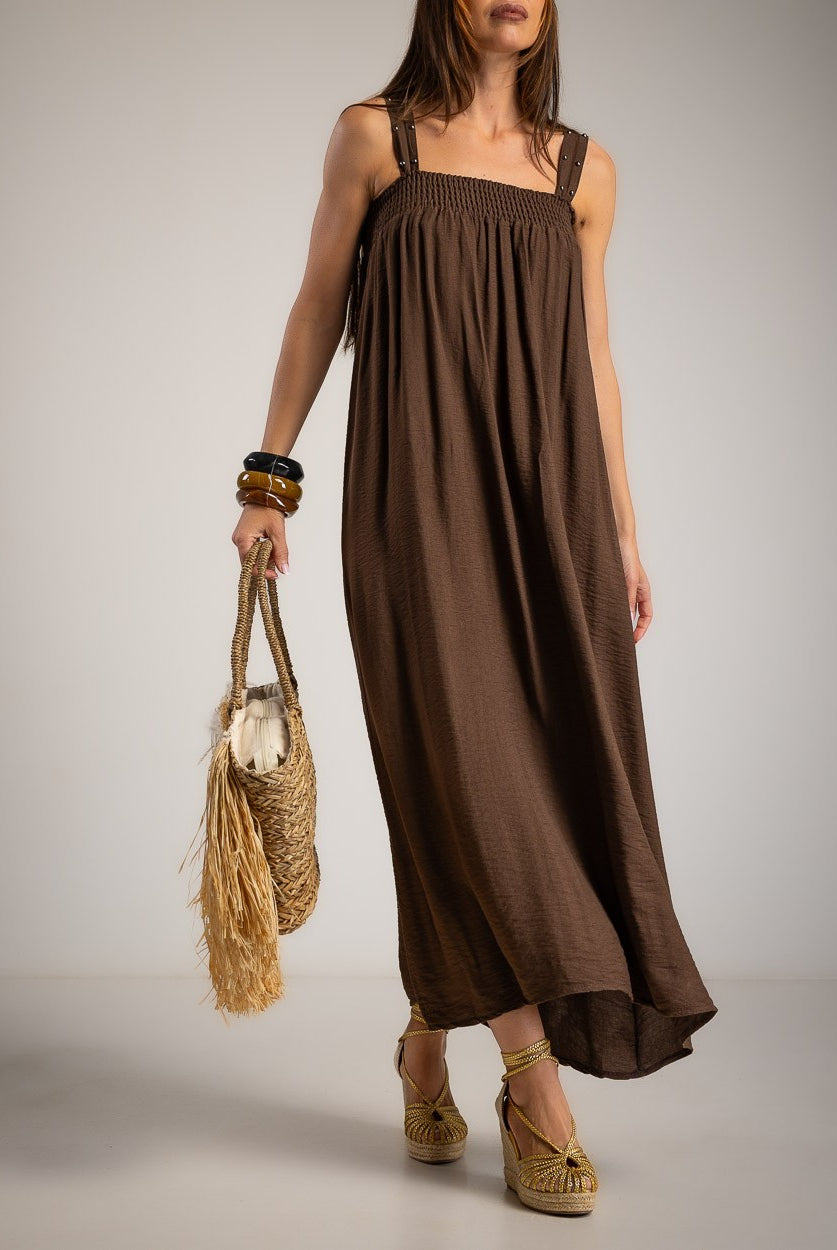 A woman models a brown sleeveless maxi dress with a smocked bodice and flowing skirt, holding a straw bag and wearing woven sandals against a neutral studio background.