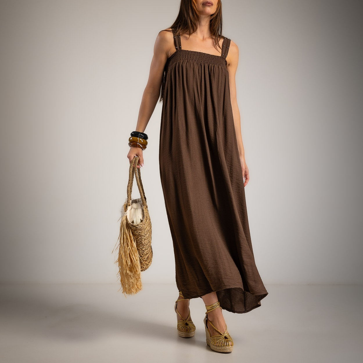 A woman models a brown sleeveless maxi dress with a smocked bodice and flowing skirt, holding a straw bag and wearing woven sandals against a neutral studio background.
