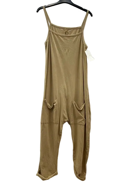 A camel sleeveless jumpsuit with thin shoulder straps, a square neckline, and two large patch pockets on the front, hanging on a black hanger against a wrinkled white fabric backdrop.