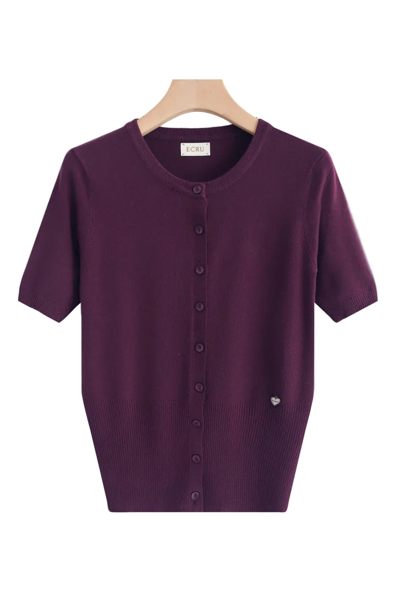 A short-sleeve knit cardigan top in deep plum purple, hanging on a light wood hanger against a clean white background. The cardigan features a round neckline, a front button closure, and a small silver heart detail on the lower right side.