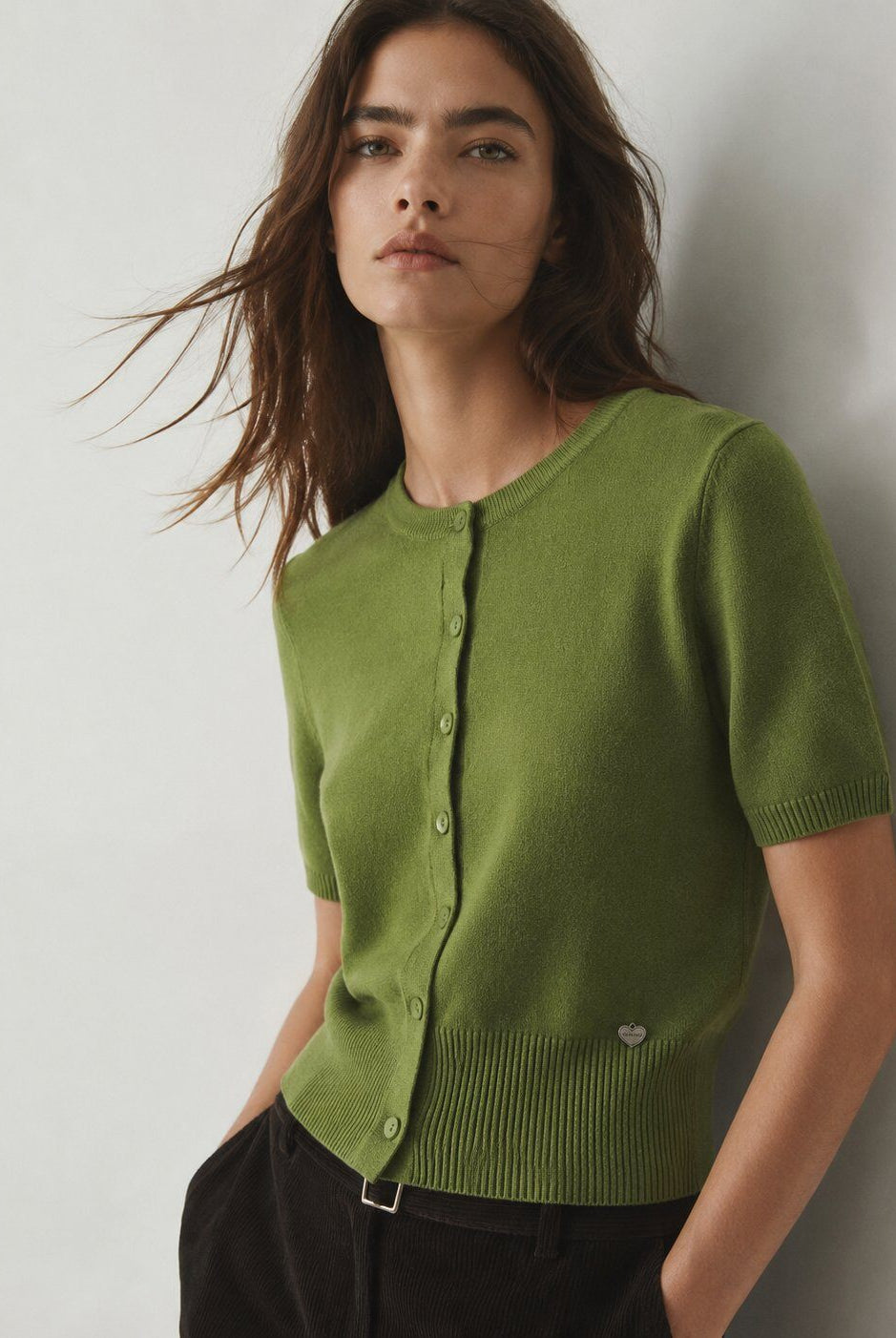 A woman wearing a short-sleeve knit cardigan in olive green with a button-front closure and ribbed hem, paired with dark pants, posed against a plain light gray wall.