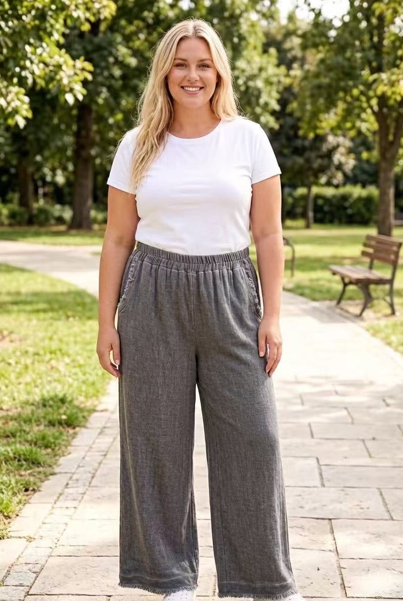Woman wearing a white t-shirt and charcoal-grey-wide-leg pants standing on a paved path with greenery in the background.
