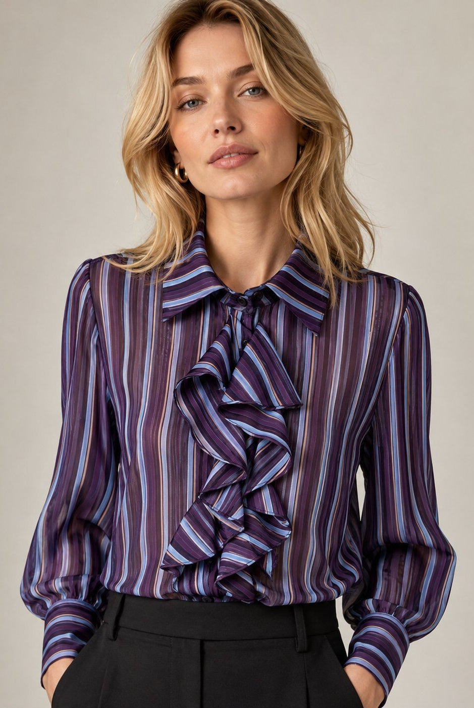 A woman wearing a sheer long-sleeve blouse with vertical purple, blue, and white stripes and a large ruffled tie-neck detail, paired with black trousers, against a neutral beige background.