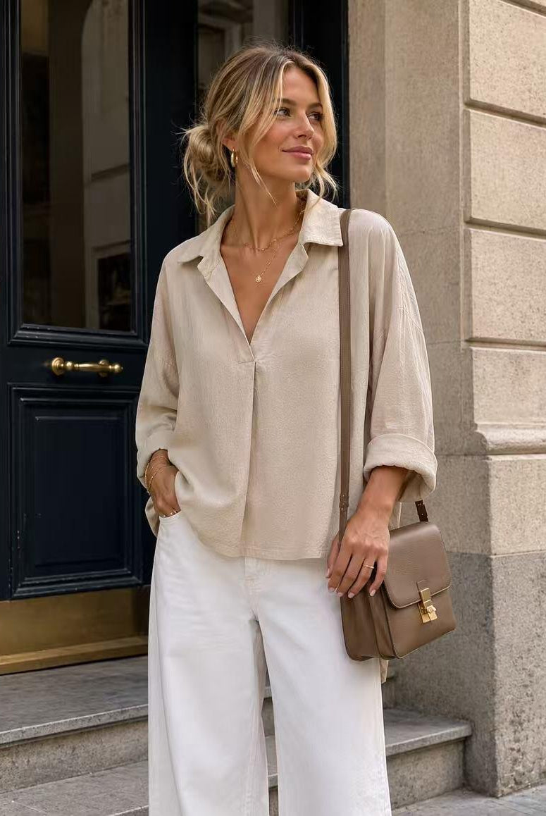 A woman stands on stone steps in front of a dark blue door, wearing a light beige linen button-up shirt, white wide-leg pants, and carrying a taupe leather shoulder bag.
