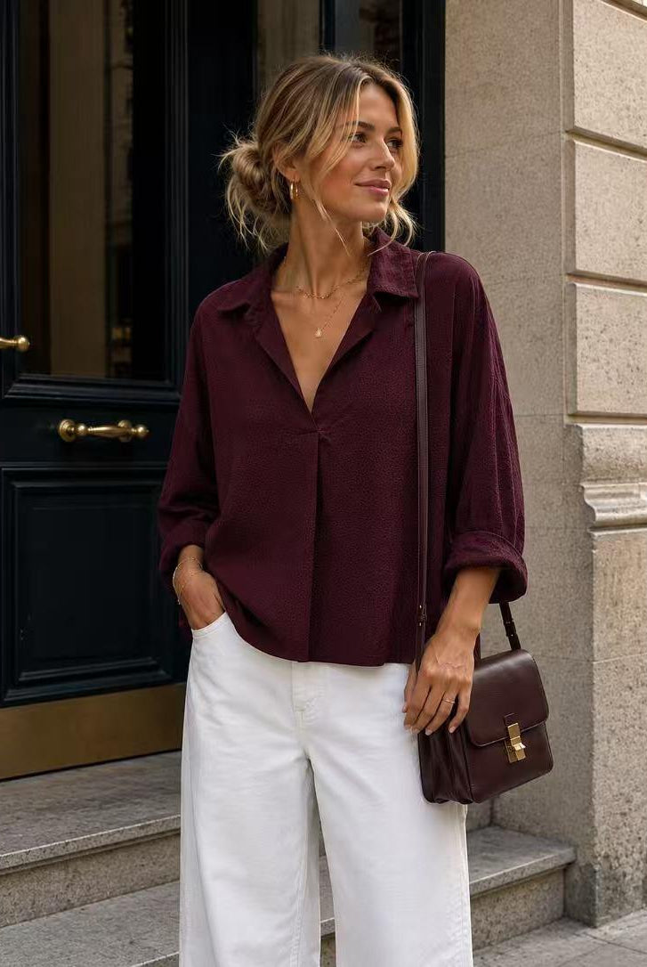 A woman stands on stone steps wearing a burgundy shirt and white wide-leg pants, carrying a matching burgundy shoulder bag.