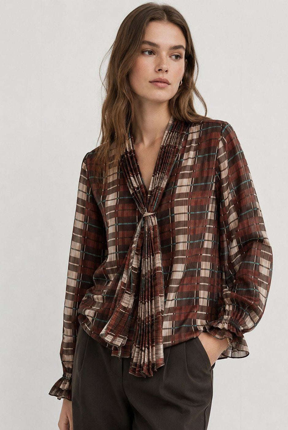 A woman modeling a long-sleeve plaid blouse with a gathered, tie-front neckline and ruffled cuffs, paired with dark trousers against a light gray studio background.