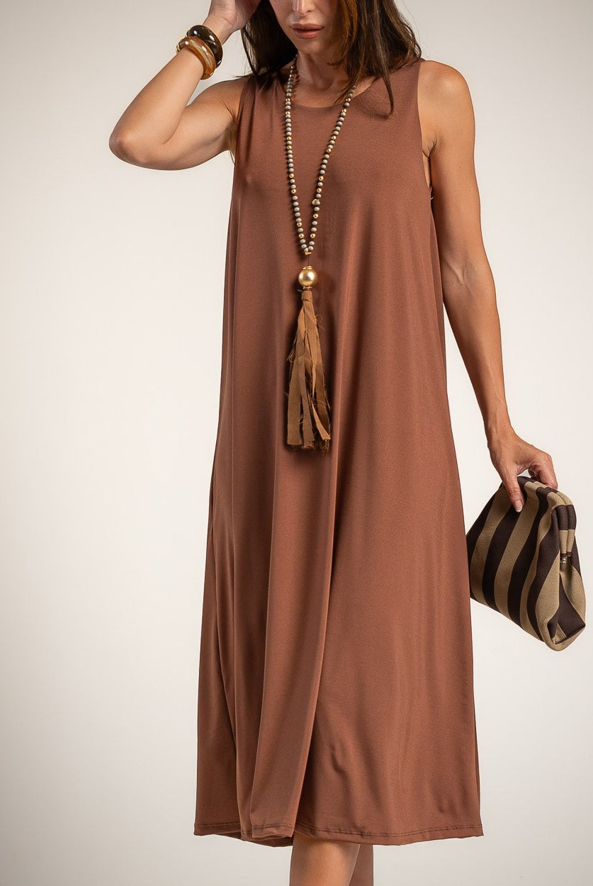 A woman wearing a sleeveless, midi-length brown dress with a tassel necklace and holding a striped clutch bag against a plain light background.