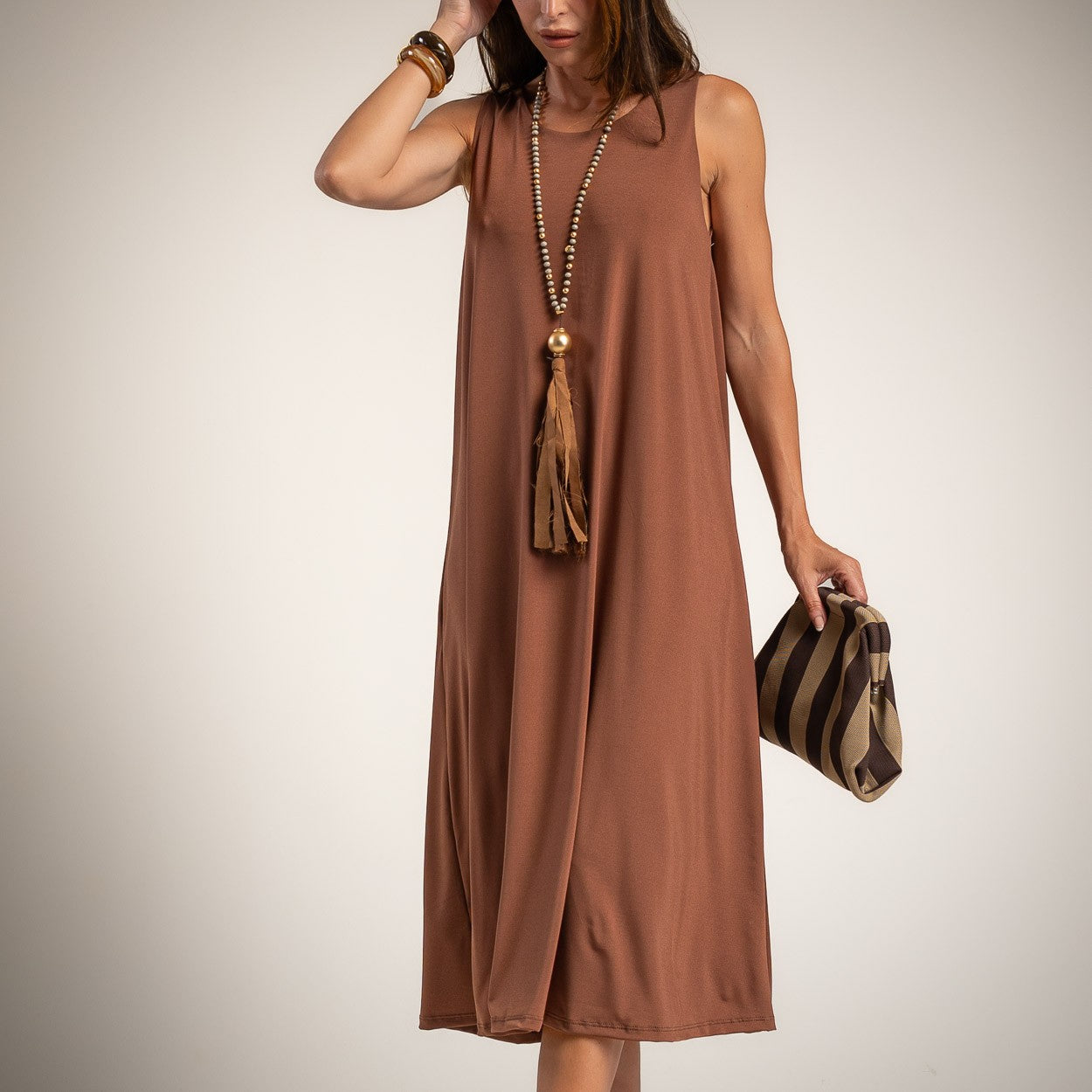 A woman wearing a sleeveless, midi-length brown dress with a tassel necklace and holding a striped clutch bag against a plain light background.