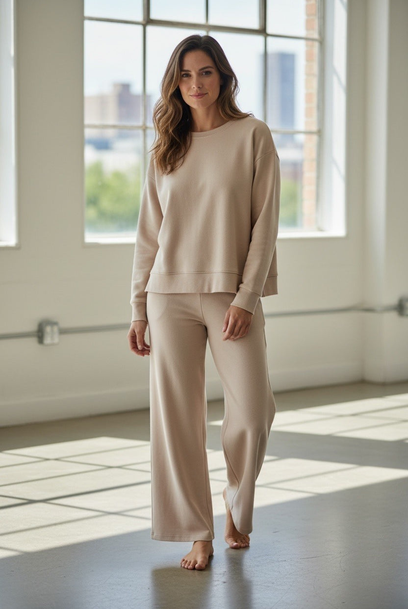 Woman wearing a beige outfit standing in a bright room with large windows.