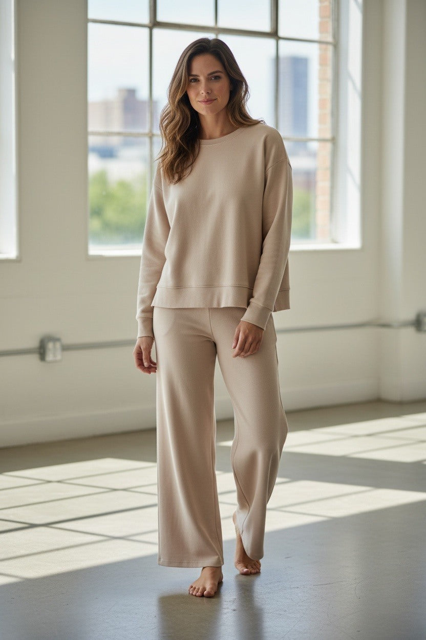 Woman wearing a beige outfit standing in a bright room with large windows.