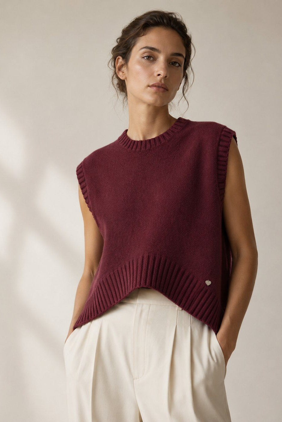 A woman wearing a sleeveless ribbed knit sweater vest in deep burgundy, paired with cream-colored trousers, standing against a warm beige wall with soft window shadows.