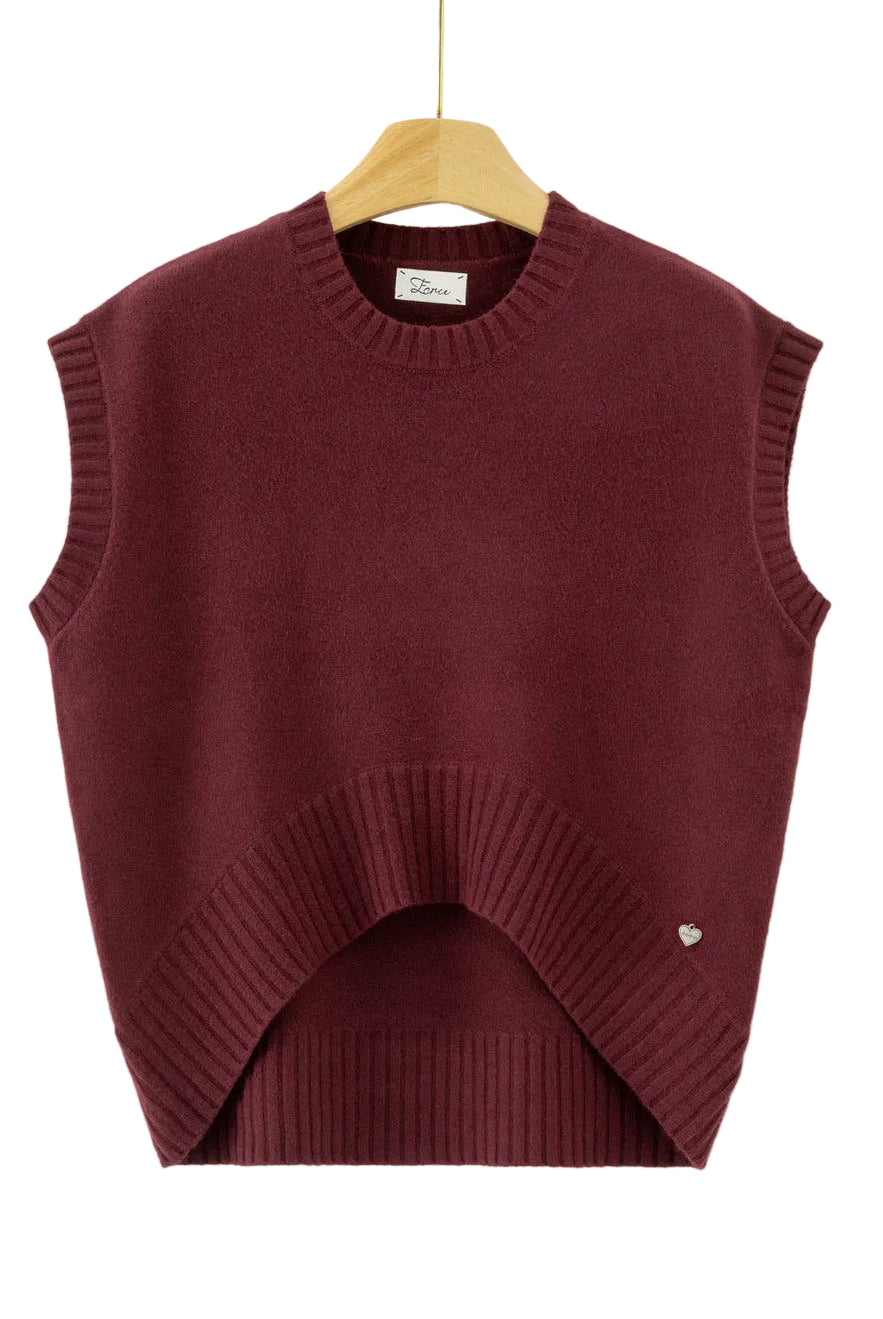 A sleeveless crewneck knit sweater vest in deep burgundy, hanging on a wooden hanger against a plain white background. The vest features a unique asymmetrical hem with a ribbed arch and a small silver heart detail on the lower right side.