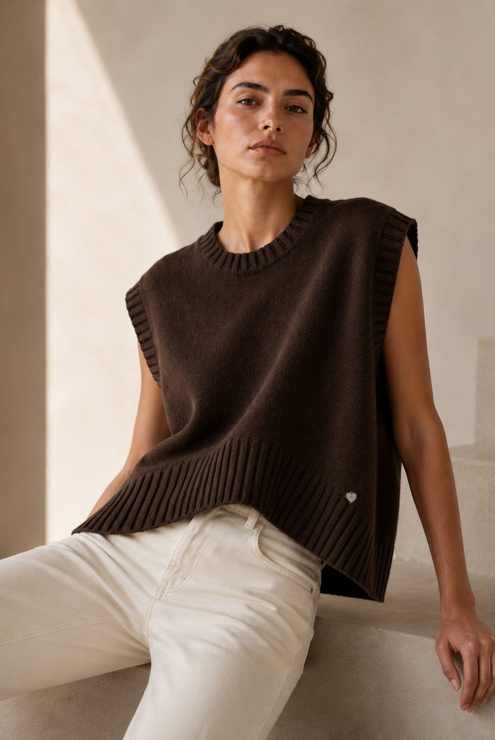 A woman wearing a sleeveless dark brown ribbed knit sweater vest and off-white trousers, seated on a light-colored stone step in a warm, minimalist interior.