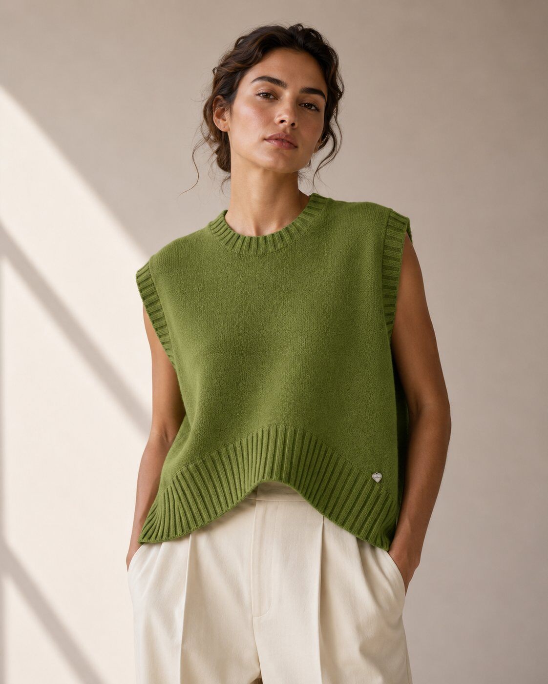 A woman wearing a sleeveless olive green knit sweater vest with ribbed trim over cream-colored trousers, standing against a neutral beige wall with soft diagonal shadows.