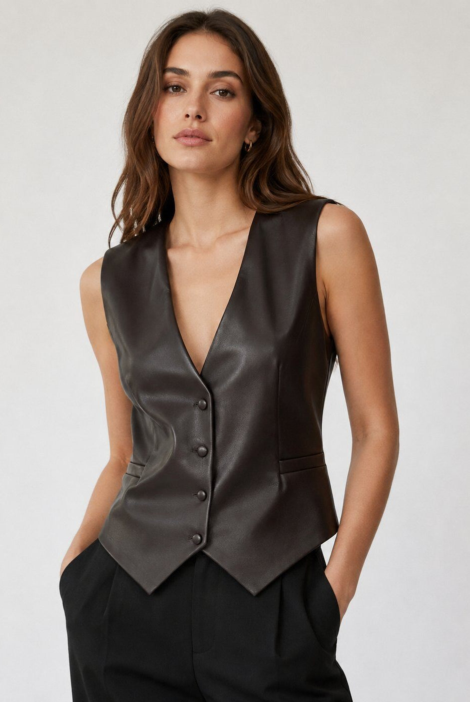 A woman modeling a dark brown leather vest with a deep V-neck and button-front closure, paired with black trousers, against a plain light gray studio background.