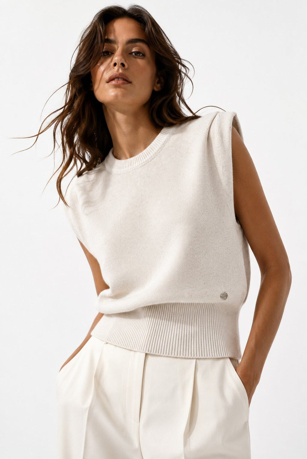 A woman wearing a sleeveless cream knit sweater vest with a ribbed hem, paired with matching cream trousers, against a clean white studio background.