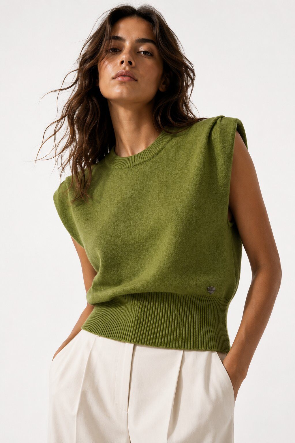 A woman wearing a sleeveless olive green knit sweater with a crew neck and ribbed hem, paired with white trousers, photographed against a clean white studio background.