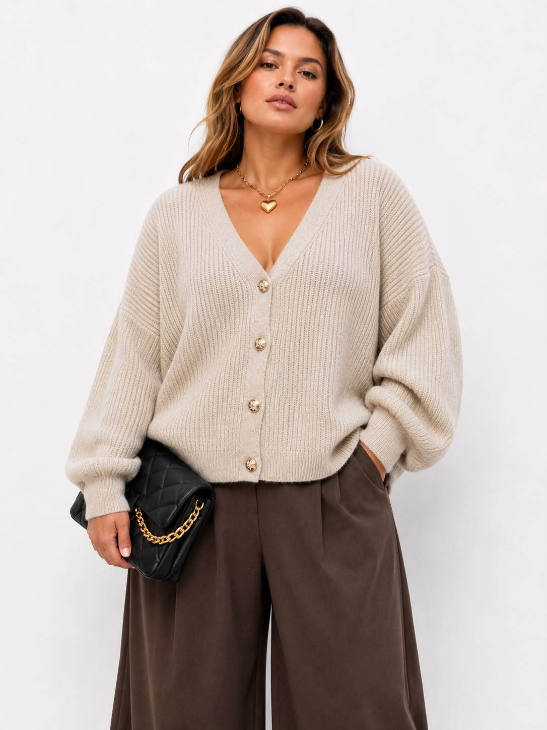 A woman wearing a beige ribbed knit cardigan with gold buttons, paired with brown wide-leg trousers and holding a black quilted clutch bag with a gold chain strap, set against a plain white background.