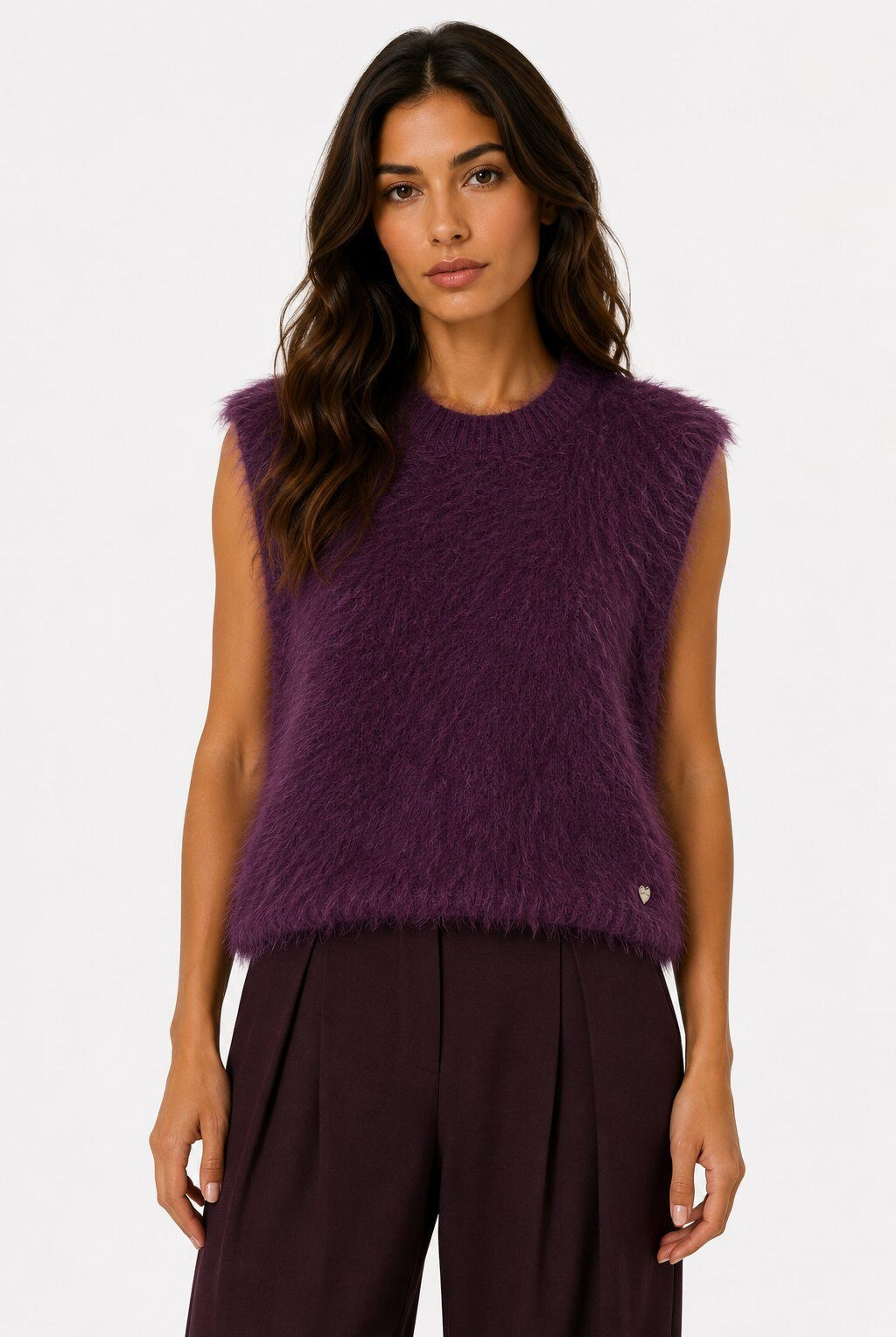 A woman modeling a sleeveless, fuzzy knit top in a deep plum purple color, paired with dark pleated trousers, against a plain white studio background.