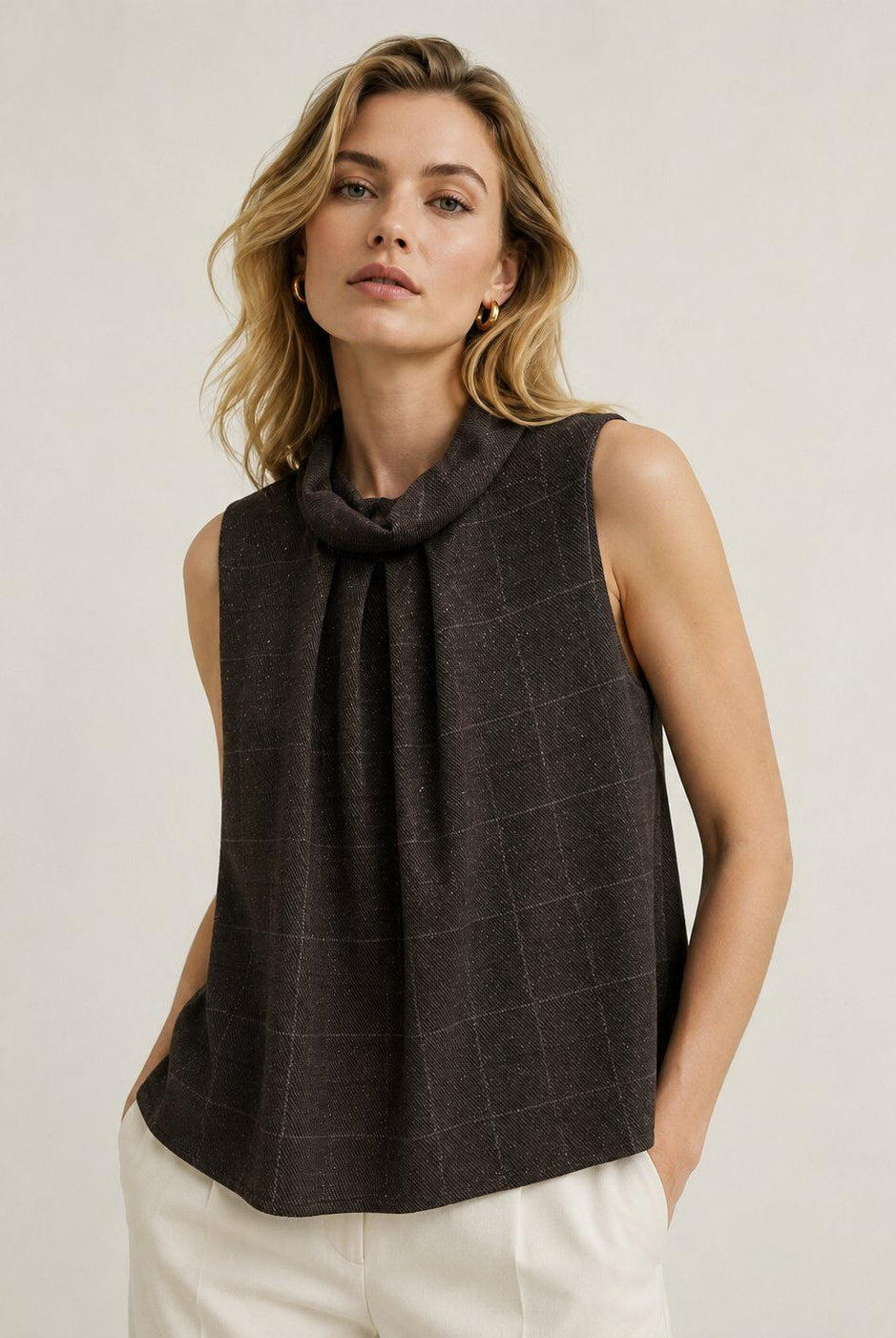 A woman wearing a dark brown sleeveless top with a cowl neck and a subtle windowpane plaid pattern, paired with off-white trousers, against a plain light beige background.