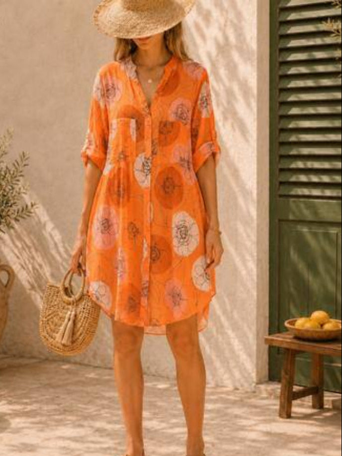 A woman wearing a short-sleeve orange button-up shirt dress with a large floral print, styled with a straw hat, woven bag, and sandals in an outdoor patio setting.