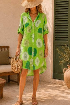 A woman wearing a bright green short-sleeve button-up dress standing in a sunlit outdoor patio setting with a woven straw hat, straw bag, and sandals.