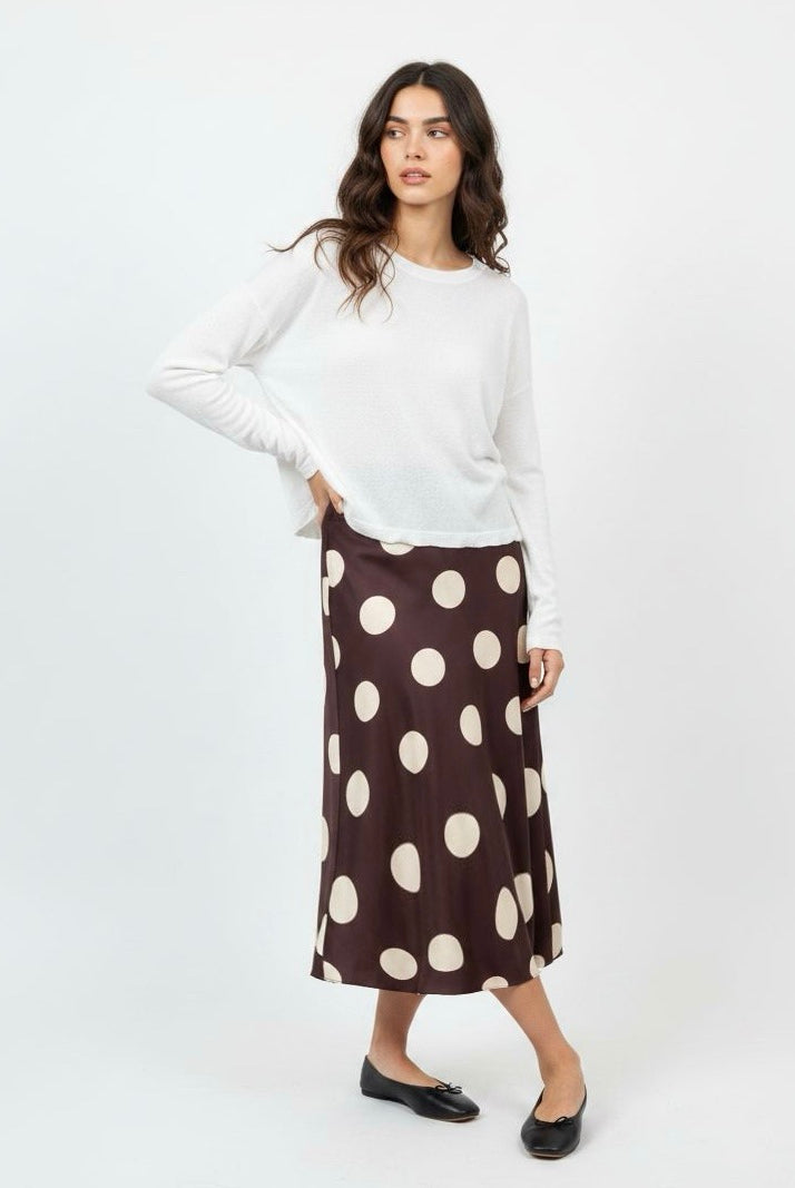 A woman modeling a white long-sleeve knit top and a brown midi skirt with large cream polka dots, paired with black flats, against a plain white studio background.